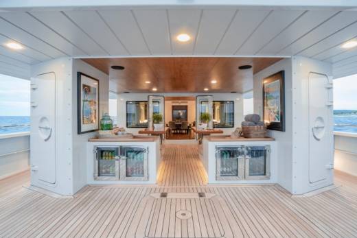 A spacious, well-lit deck with wooden flooring in the foreground leads to a bar area with refrigerators and artwork, opening into a dining area with mirrors and tables in the background.