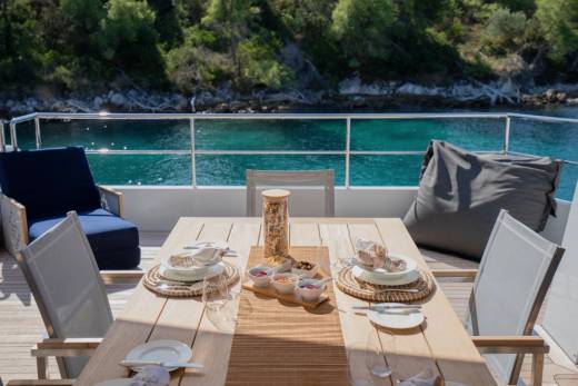 A dining table set for a meal on a yacht's deck overlooks a tranquil turquoise sea and lush green hills.