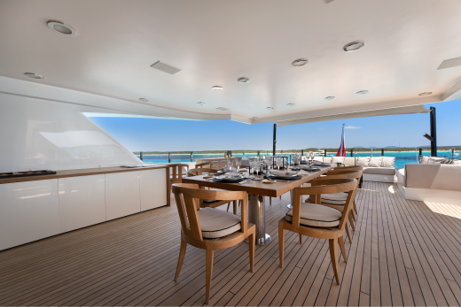 A long dining table set for a meal sits on a wooden deck in the foreground, with a bright blue ocean and distant islands visible in the background.