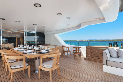 A wooden deck with a dining table set for a meal in the foreground leads to a bright blue ocean and coastline in the background.