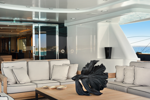 A modern yacht's outdoor lounge area with plush white sofas and a striking black sculpture in the foreground, opens to a bright blue ocean and sky in the background.