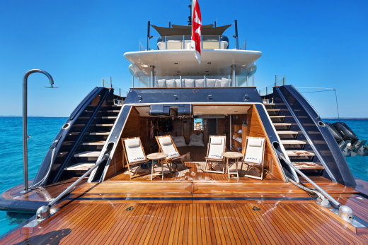 A luxurious yacht with a spacious deck and lounge area sits on a clear turquoise sea under a bright blue sky.