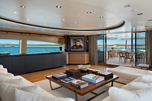 A luxurious yacht interior with a plush white sofa and coffee table in the foreground, and a scenic ocean view with an outdoor dining area in the background.