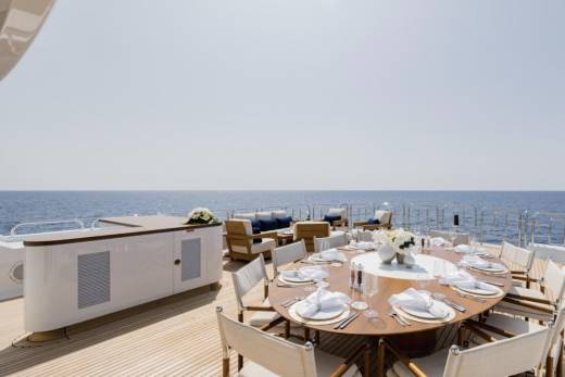 A large oval dining table set for a meal on a yacht's deck is surrounded by chairs, with the calm blue ocean stretching to the horizon in the background.