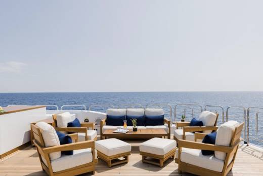 A luxurious outdoor seating area with white cushions and navy accents is arranged on a wooden deck, with the calm blue ocean and clear sky stretching out in the background.