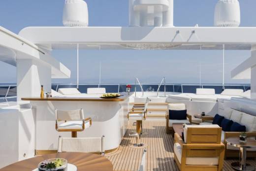 A luxurious yacht deck with a bar and seating in the foreground, and the calm blue ocean and sky in the background.