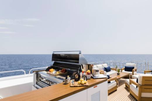 A stainless steel grill and a wooden bar are in the foreground, with a seating area and the ocean stretching to the horizon in the background.