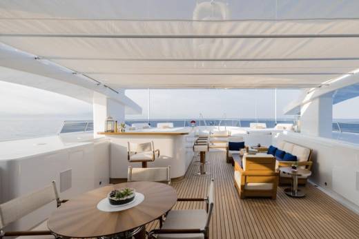 A circular wooden table with chairs in the foreground is set on the deck of a yacht, with a bar and lounge area visible in the background against a backdrop of the ocean and sky.