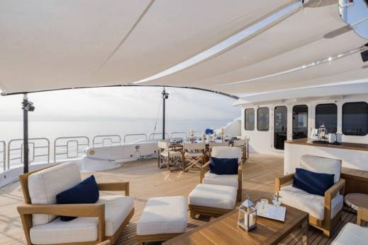 In the foreground, comfortable white lounge chairs with blue cushions are arranged on a wooden deck, while the background reveals a serene ocean view under a large white canopy.