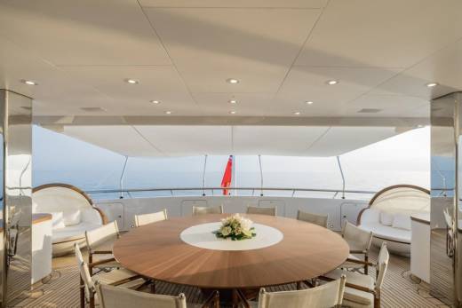 A large, round wooden dining table with chairs is set on a yacht's deck, with the calm ocean and a red flag visible in the background.