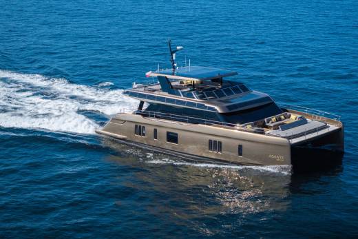 A sleek, golden catamaran yacht glides through calm blue water, leaving a white wake behind it, with a clear sky visible in the distance.