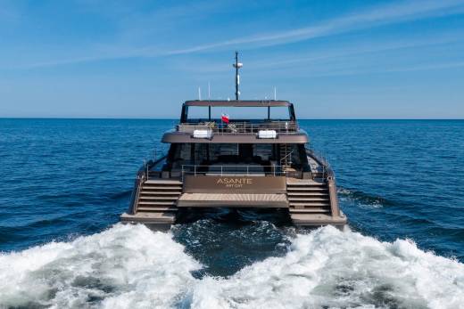A modern catamaran yacht named "ASANTE" cruises through calm blue waters, leaving a white wake behind it under a clear sky.