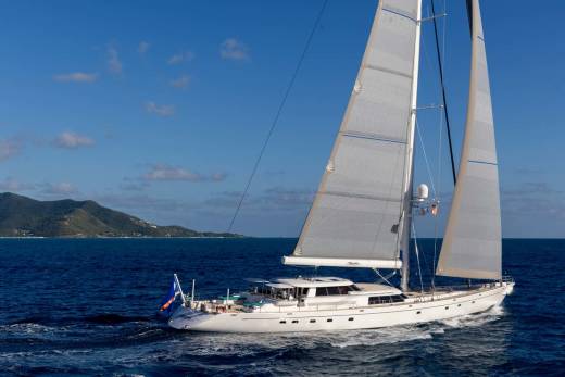 A large white sailboat with full sails glides across a deep blue ocean, with a lush green island visible in the hazy background under a clear blue sky.