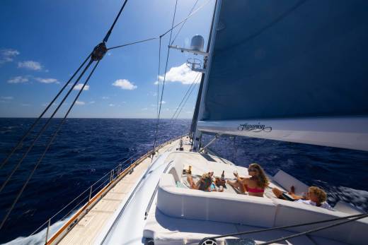 People relax on the deck of a sailboat with a blue sail against a bright blue sky and ocean.