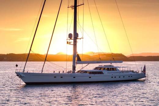 A large white sailboat rests on calm water in the foreground, silhouetted against a vibrant orange sunset over distant hills in the background.