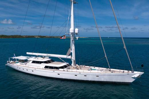 A large white yacht with tall masts sails on a calm blue sea, with a distant, hazy coastline under a bright sky.