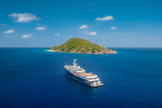 A large yacht sails on deep blue water in the foreground, with a lush green island in the background under a bright blue sky dotted with white clouds.