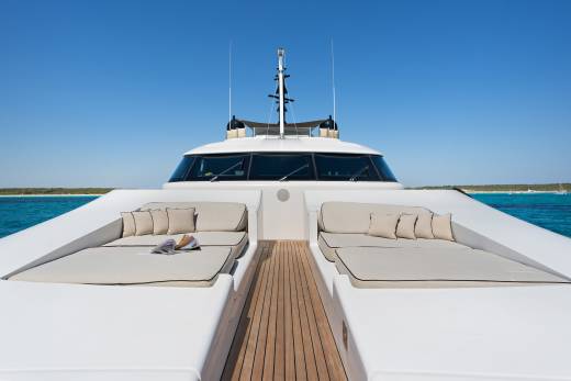 Two lounge areas with beige cushions and pillows sit on the white deck of a yacht, with a clear blue sky and ocean in the background.