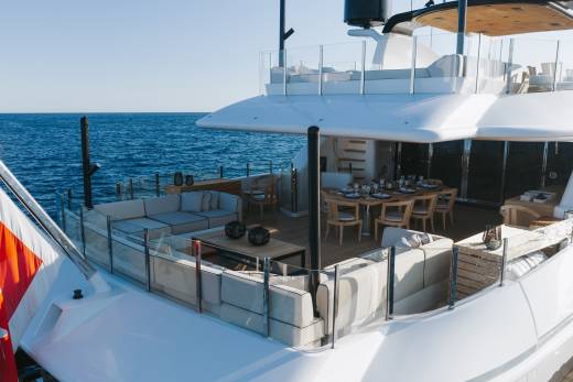 A luxury yacht deck features white lounge seating and a dining area in the foreground, set against a backdrop of a deep blue ocean under a clear sky.