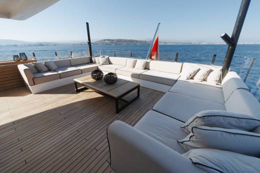 A luxurious yacht deck features white sectional sofas and a wooden coffee table set against a backdrop of a calm blue ocean and distant hills.