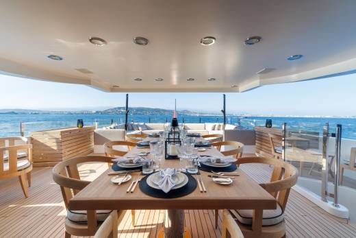 A set dining table with wooden chairs sits on the deck of a luxury yacht, overlooking a calm blue sea and distant coastline.