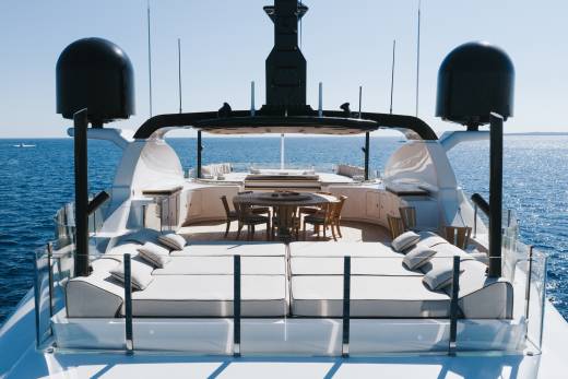 A luxury yacht deck features white lounge seating and a wooden dining area in the foreground, set against a vast blue ocean and clear sky in the background.