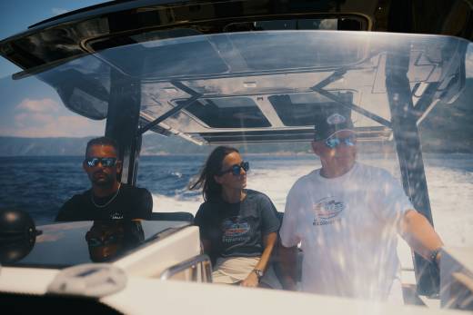 Three people wearing sunglasses sit inside a boat's cabin with a vast blue ocean and distant coastline in the background.