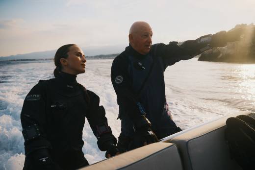 A man and woman in wetsuits stand on a boat, with the man pointing toward a distant coastline across the churning ocean water.