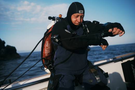 A scuba diver in a black hood and blue wetsuit prepares his gear on a boat against a backdrop of blue ocean and sky.