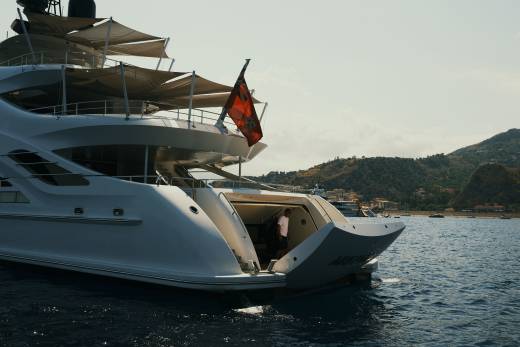 A large white luxury yacht with a red flag floats in the foreground against a backdrop of calm blue water and distant green mountains.