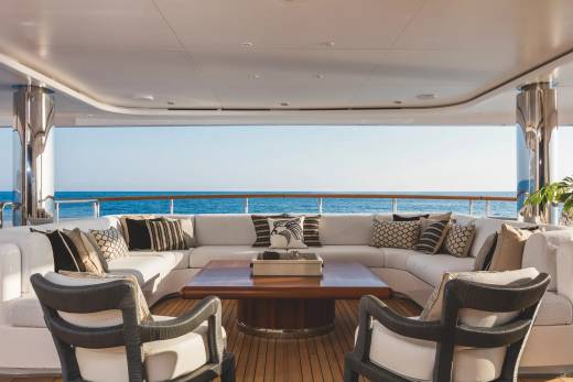 A luxurious white sectional sofa and two armchairs surround a wooden coffee table on a yacht deck, overlooking a calm blue ocean under a clear sky.