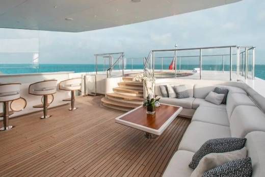 A luxurious yacht deck features a white lounge area with a wooden coffee table and bar stools in the foreground, overlooking a calm blue ocean in the background.