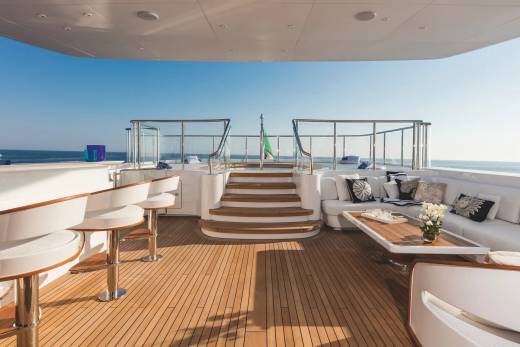 A luxury yacht deck features white seating and a wooden floor in the foreground, leading to stairs and a railing with a clear blue ocean and sky in the background.