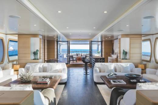 A luxurious yacht interior features white sofas and wooden coffee tables in the foreground, leading toward an open deck with a view of the blue ocean in the background.