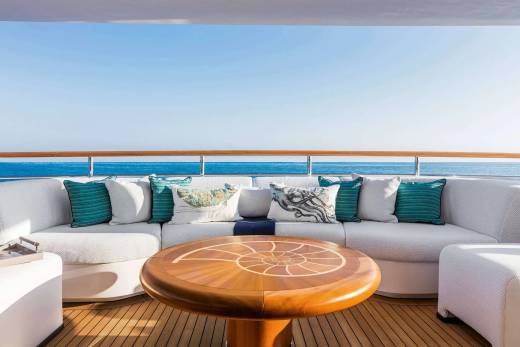 A round wooden table and a white curved sofa with teal pillows sit on a wooden deck overlooking a calm blue ocean under a clear sky.
