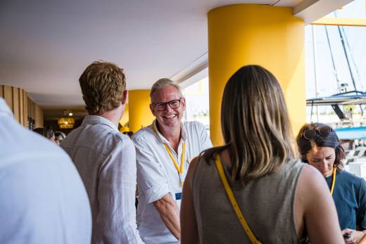 A smiling man in a white shirt stands among a group of people in the foreground, with a bright yellow pillar and a glimpse of a boat in the background.