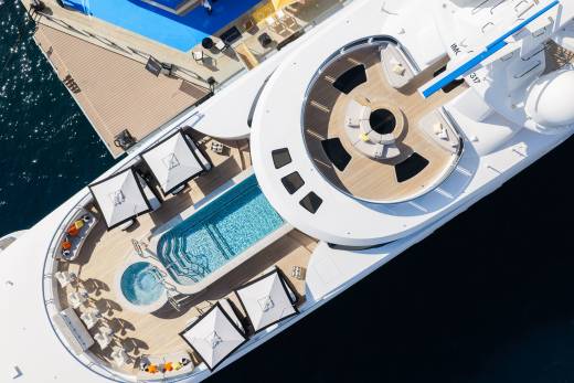 A luxury white yacht featuring a swimming pool and lounge areas sits docked next to a wooden pier and deep blue water.