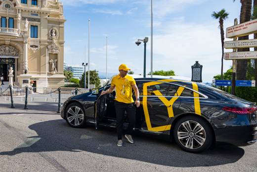 A man in a yellow shirt and cap steps out of a black car with yellow graphics, parked in front of an ornate classical building.