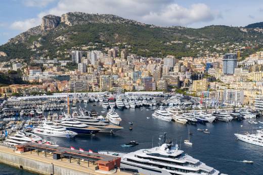 Numerous luxury yachts fill a bustling harbor in the foreground, set against a backdrop of a densely populated hillside city and towering green mountains.