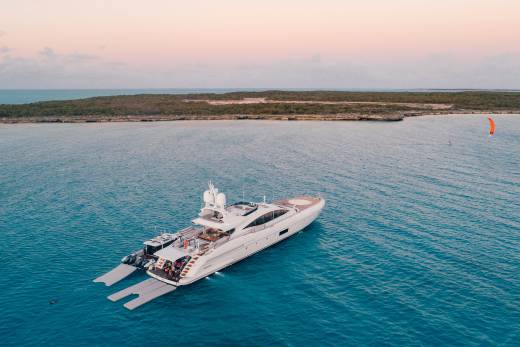 A large white luxury yacht floats in turquoise water with a scrubby coastline and a pale pink sky in the background.