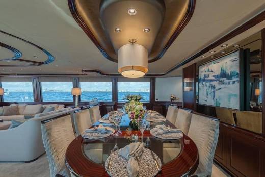 An elegant dining table set for a meal occupies the foreground of a luxury yacht interior, with a plush lounge area and ocean views visible in the background.