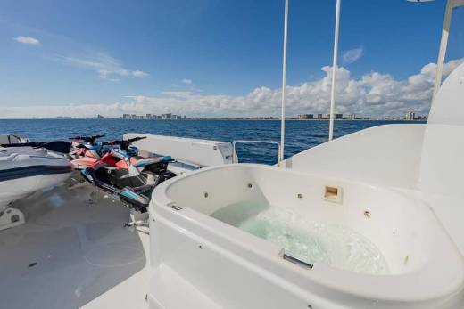 A white hot tub and jet skis sit on the deck of a boat overlooking a blue ocean and a distant city skyline under a sunny sky.