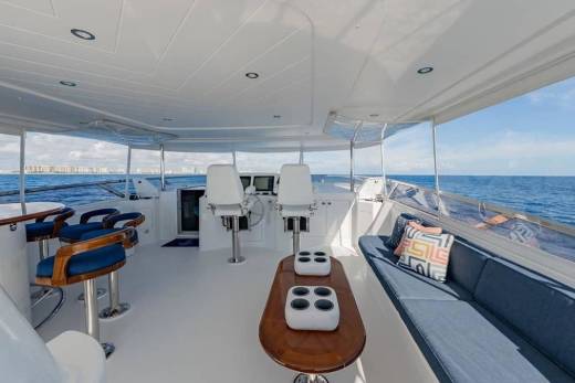 A luxurious yacht deck features a lounge area with a blue sofa and bar stools in the foreground, overlooking a calm blue ocean and a distant city skyline in the background.