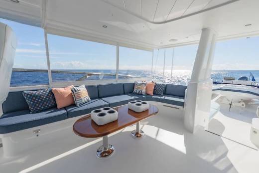A blue cushioned lounge area with a wooden coffee table sits in the foreground of a yacht deck, overlooking a sparkling blue ocean and a small boat in the background.