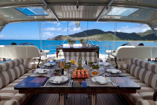 A set dining table with fruit and appetizers sits in the foreground of a luxury yacht, overlooking a turquoise ocean and lush green mountains in the background.