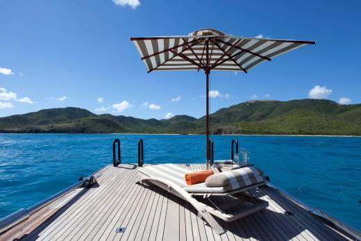 A striped beach umbrella and lounge chair sit on the wooden deck of a boat, overlooking a bright blue ocean and lush green mountains.