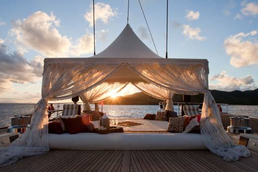 A luxurious white canopy bed with plush pillows sits on a wooden deck, set against a backdrop of calm ocean waters and distant mountains during a golden sunset.