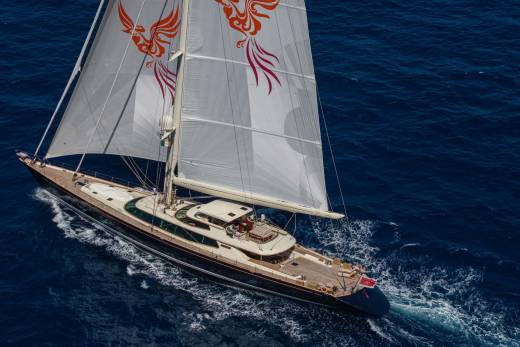 A large sailing yacht with white sails featuring orange phoenix emblems glides through deep blue ocean waters.