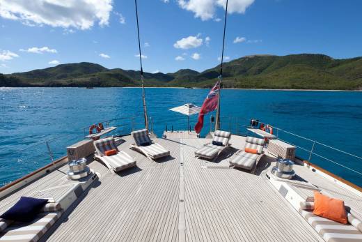 A wooden yacht deck with lounge chairs and a red flag sits in the foreground, set against a backdrop of turquoise water and lush green mountains under a blue sky.