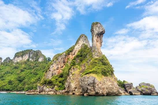 A towering, chicken-shaped rock formation covered in lush green vegetation rises from the turquoise sea, with more verdant islands and a bright blue sky with scattered clouds in the background.